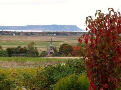 A bright red tree and wildflowers stand in the foreground of this autumn photo taken from a look off in Grand Pré, Nova Scotia. Beyond is the Grand Pre National Historic Park and the church of the Acadian people. In the background is Cape Blomidon and the Bay of Fundy in the beautiful Annapolis Valley