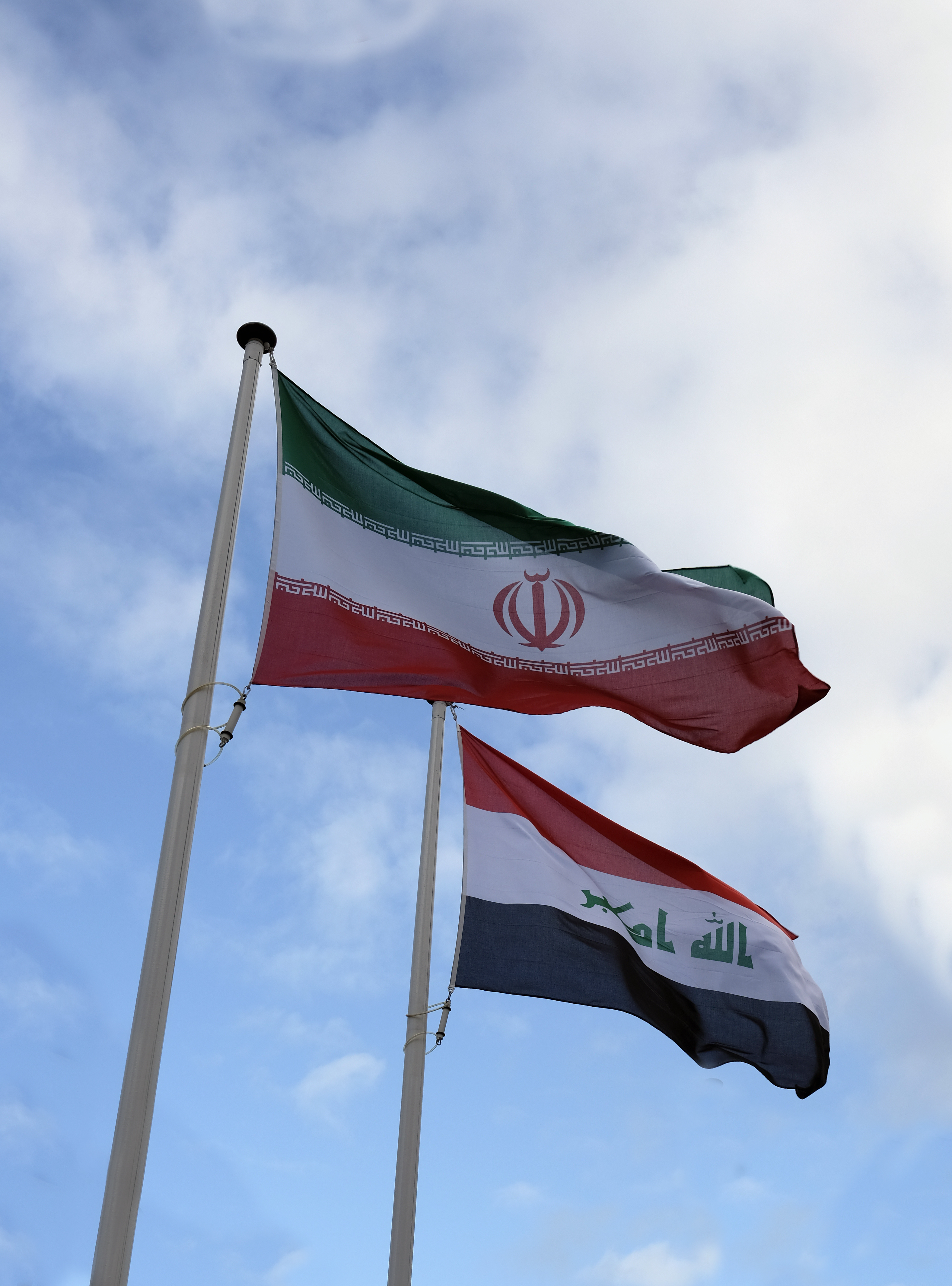 Les drapeaux iranien et irakien flottent devant un ciel bleu partiellement ennuagé.