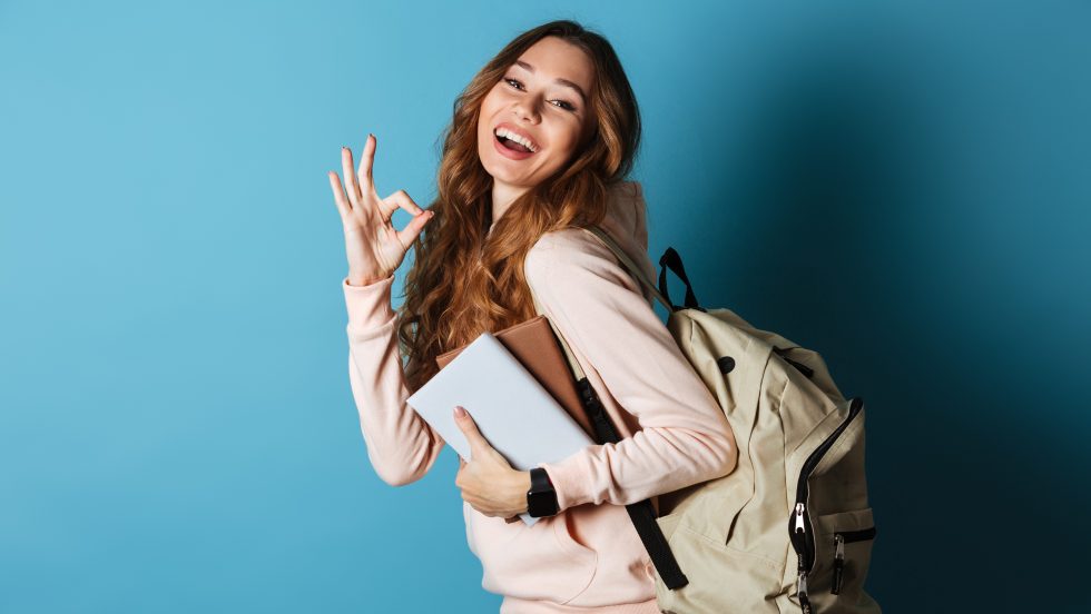 Portrait d'une étudiante heureuse avec sac à dos, tenant des livres et montrant le geste ok isolé sur fond bleu