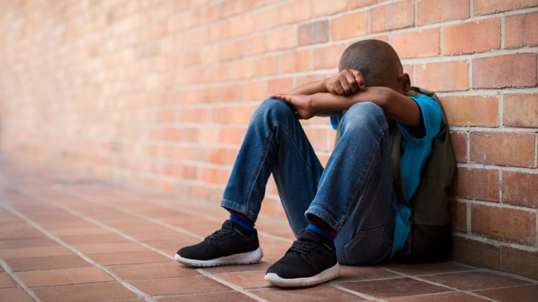 Un garçon noir se cache la figure, accroupi devant le mur de brique de son école après un incident de racisme en classe.
