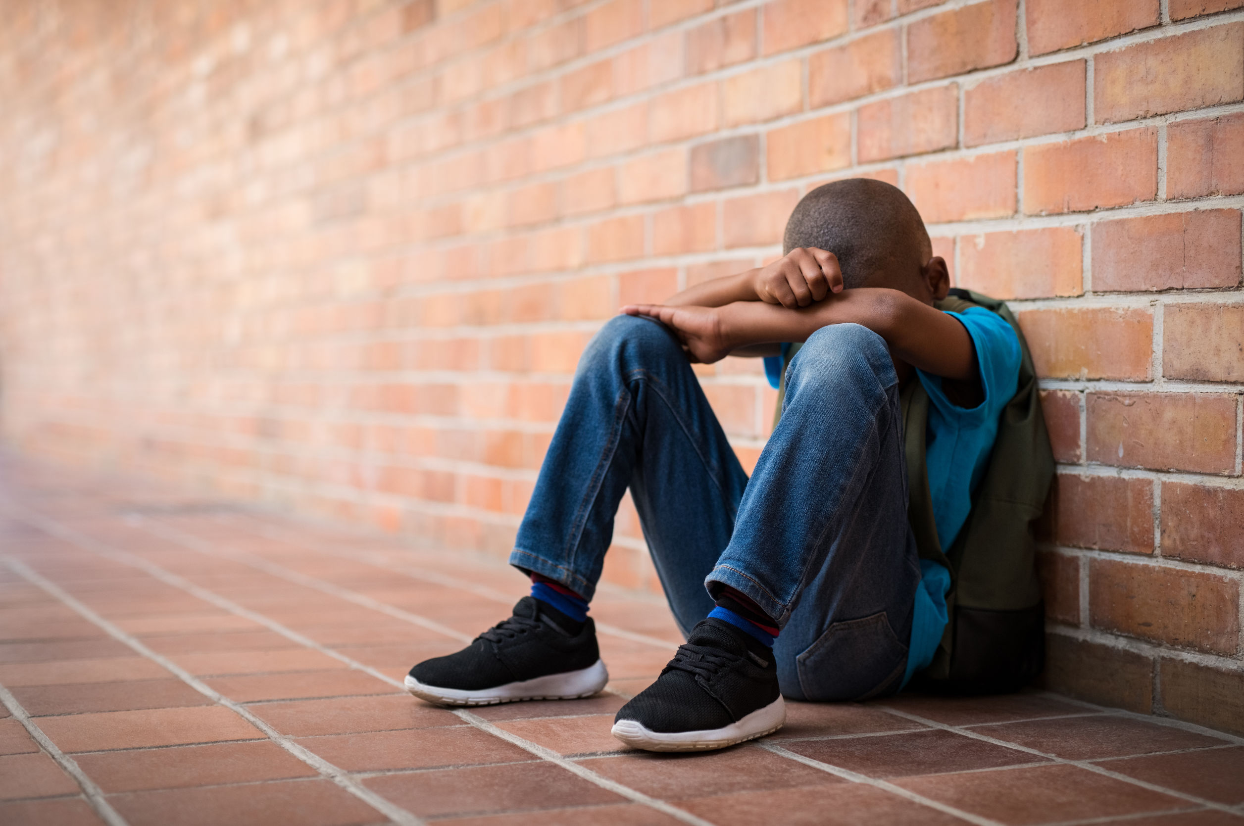 Un garçon noir se cache la figure, accroupi devant le mur de brique de son école après un incident de racisme en classe.