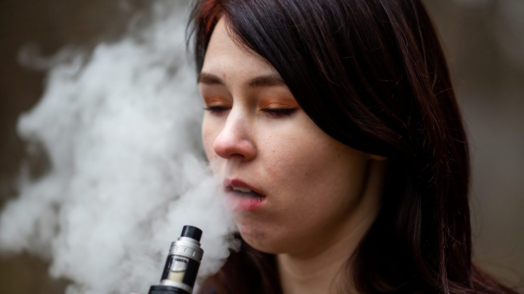 Vape teenager. Young pretty caucasian brunette girl smoking an electronic cigarette on the street in the spring. Deadly bad habit.