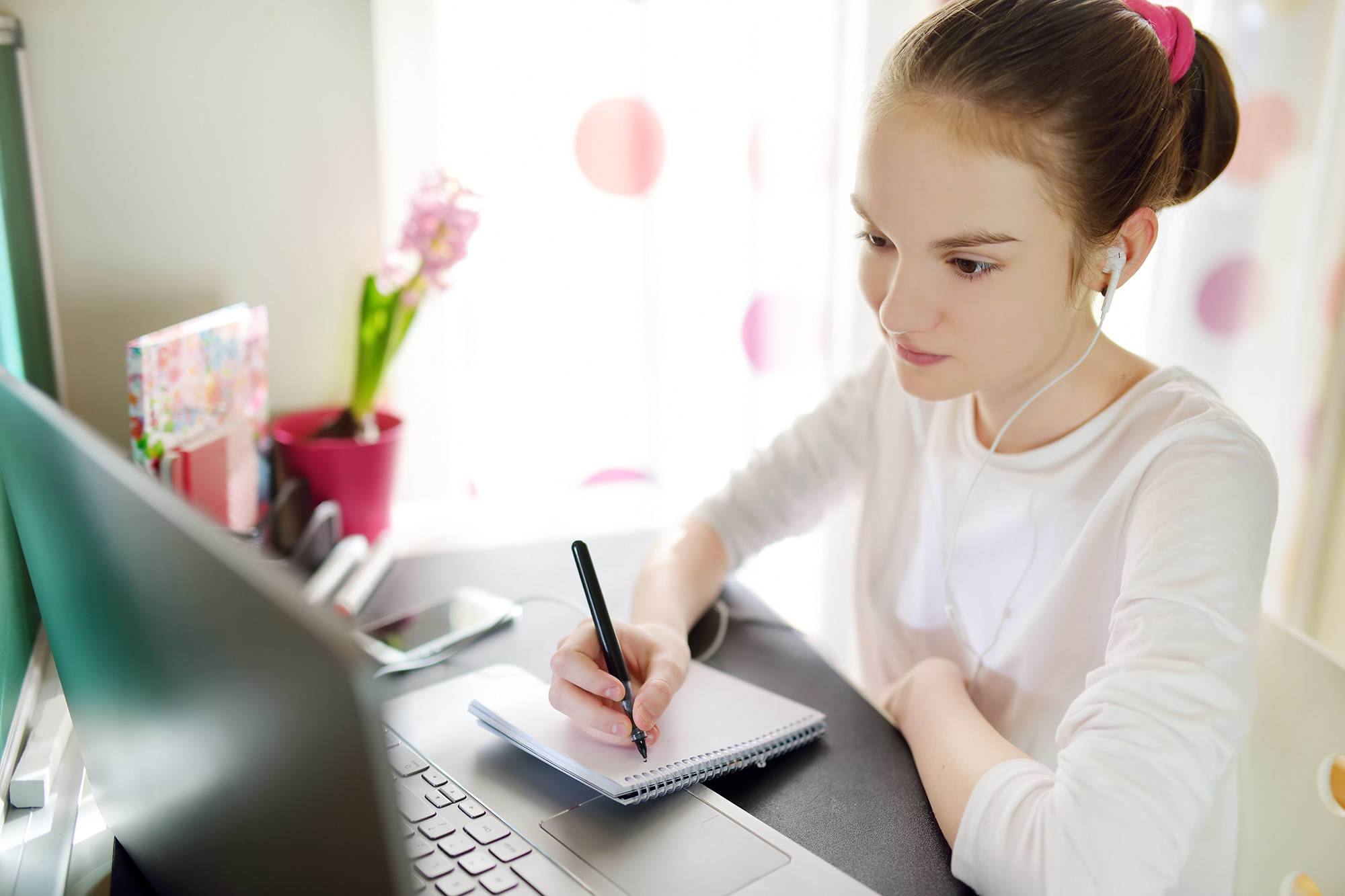 Jeune étudiante suivant un cours en ligne devant son ordinateur et prenant des notes sur un calepin.