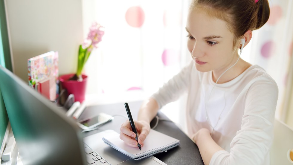 Jeune étudiante suivant un cours en ligne devant son ordinateur et prenant des notes sur un calepin.