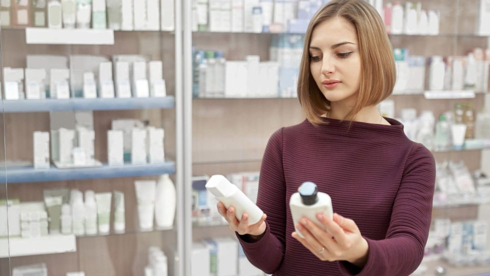 Pharmacy customer standing in drugstore and choosing medical products. Woman looking at cosmetic white bottles. Consumer buying treatment for health care.