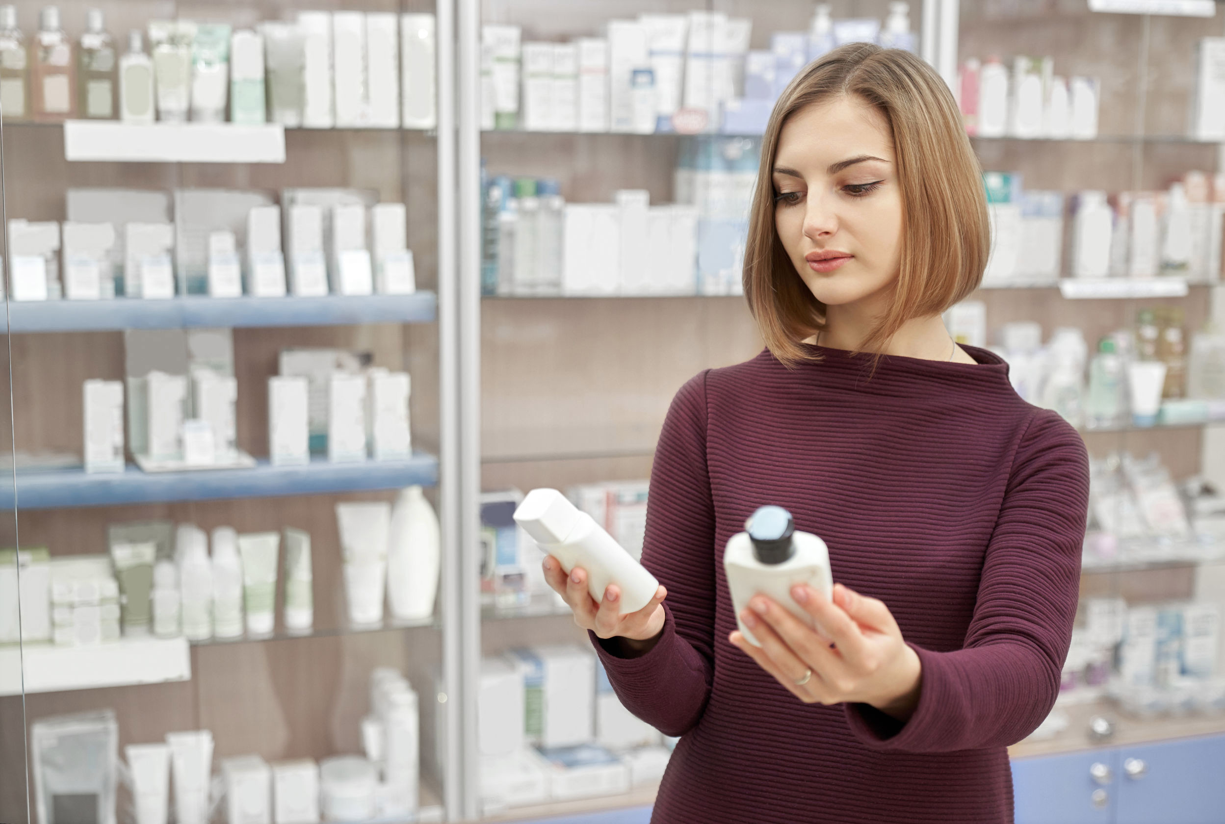 Pharmacy customer standing in drugstore and choosing medical products. Woman looking at cosmetic white bottles. Consumer buying treatment for health care.