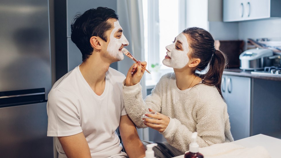 Woman applying clay mask on her boyfriends face. Young loving couple taking care of skin at home