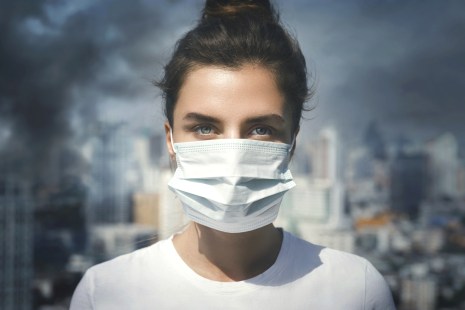 Une jeune femme portant un masque devant un environnement urbain marqué par le smog, la pollution et les gaz à effet de serre