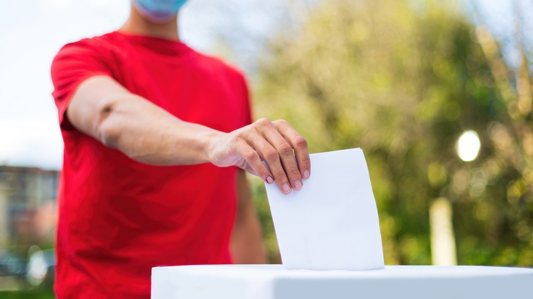 Un homme en chandail rouge portant un masque chirurgical dépose son bulletin de vote dans une boîte de scrutin, à l'extérieur devant un paysage naturel.