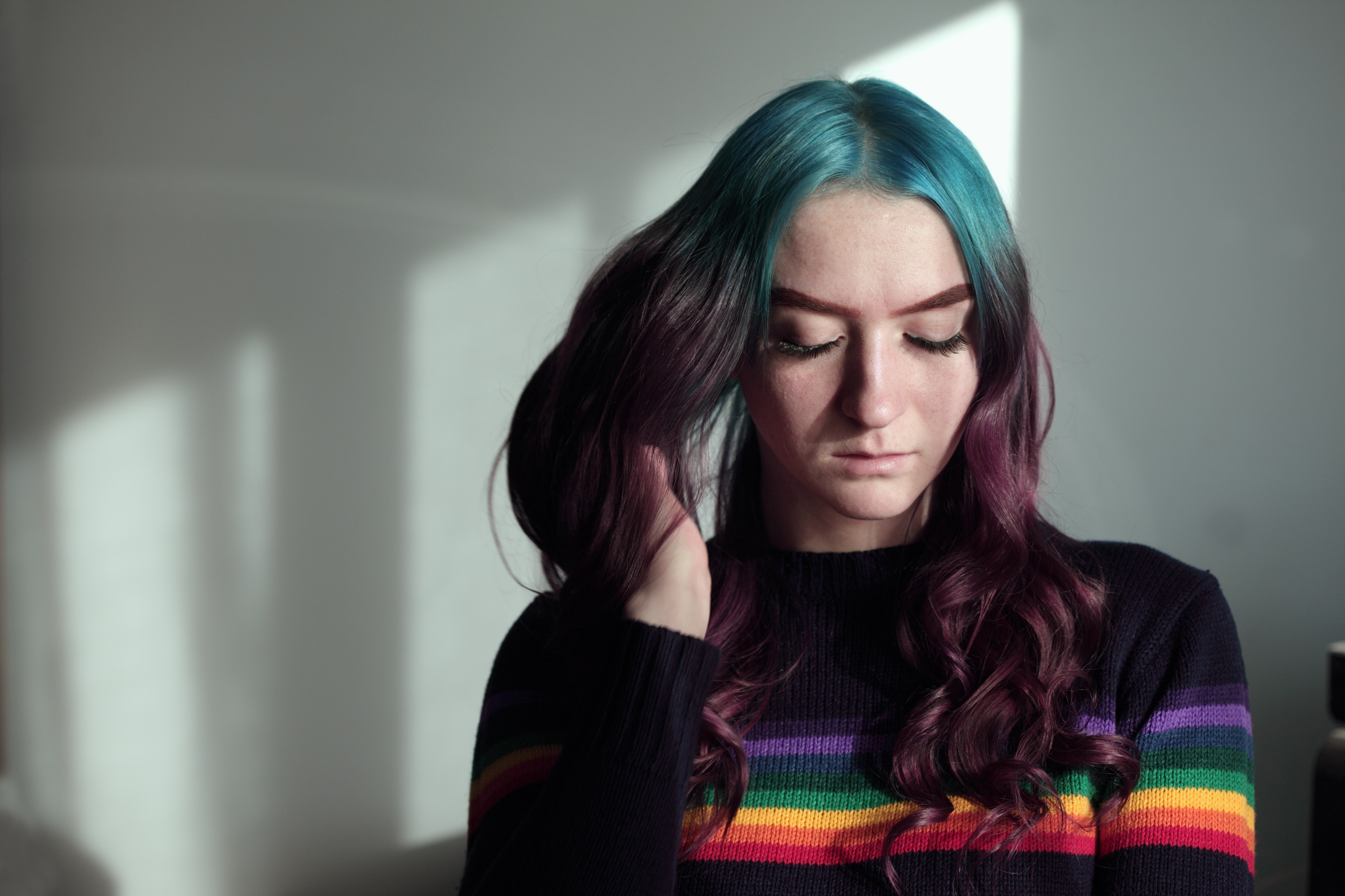 Une jeune femme avec un chandail à l'effigie de la communauté LGBTQ et des cheveux mauves et roses le regard baissé.