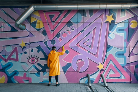 artiste de graffiti portant un manteau jaune et un masque, devant une murale