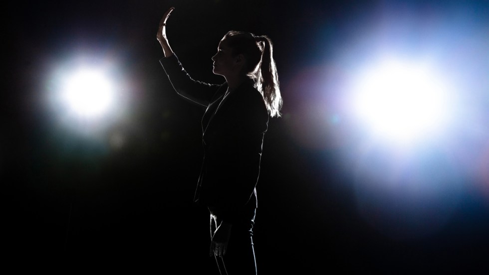 Silhouette d'une femme politicienne sur une scène, devant des projecteurs, avec une main levée.