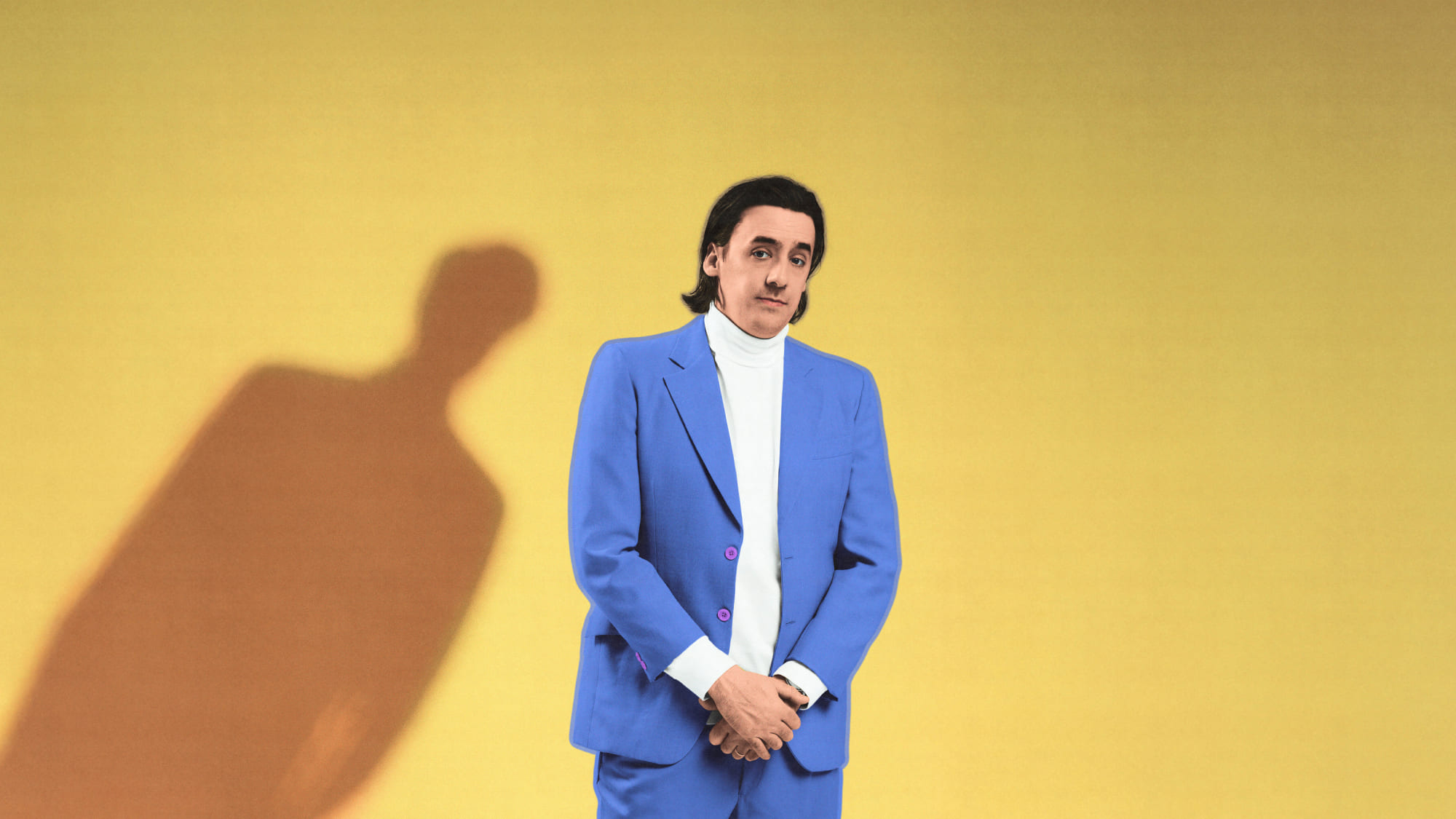 Un homme vêtu d'un complet bleu devant un fond jaune. L'ombre d'une silhouette est penchée vers l'homme. L'image illustre la pièce de théâtre «Merci d'être venus».