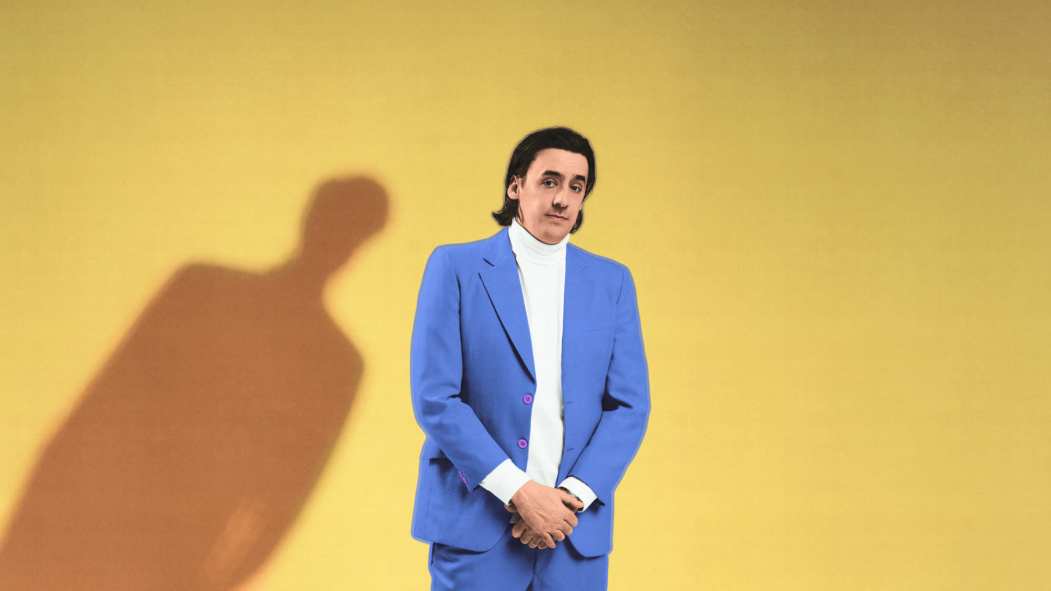 Un homme vêtu d'un complet bleu devant un fond jaune. L'ombre d'une silhouette est penchée vers l'homme. L'image illustre la pièce de théâtre «Merci d'être venus».