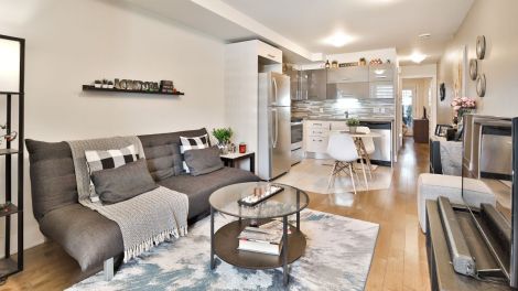 Un salon de condo meubl&eacute; d'un divan, d'une table et d'un meuble &agrave; t&eacute;l&eacute;viseur, avec en arri&egrave;re-plan une vue de la cuisine.