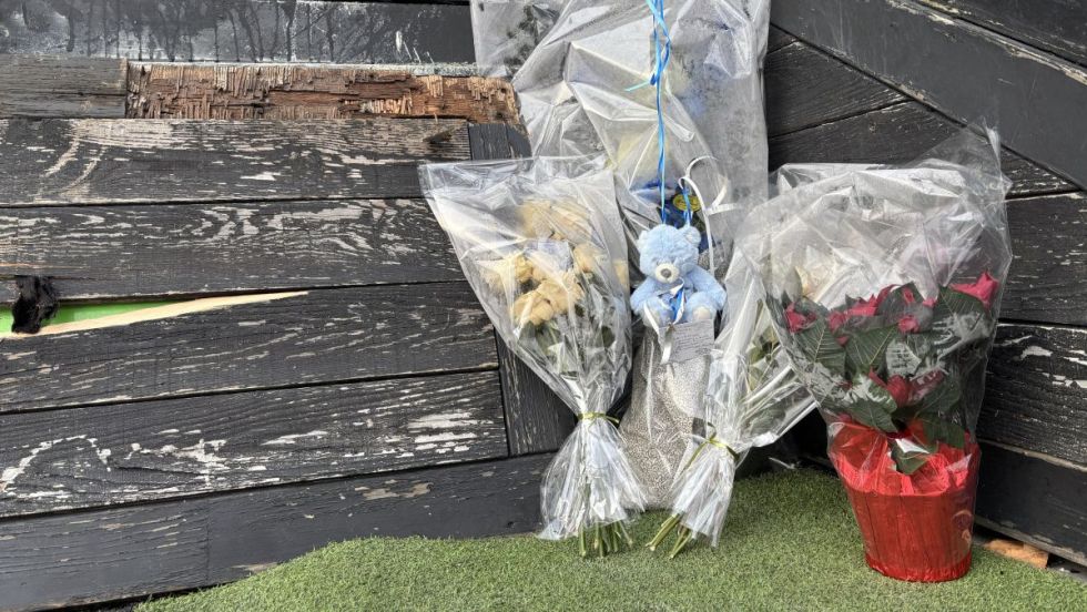 Des fleurs et un ourson en peluche sont déposées devant une fenêtre de sous-sol placardée où une personne en situation d'itinérance est décédée.