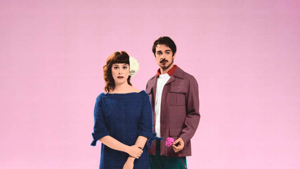 Deux personnages de la pièce Carmen, Requiem sont debout devant un fond rose. Un des personnages tient une rose dans sa main.