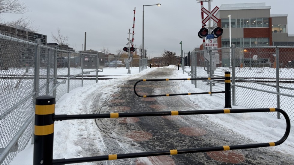 Une barrière à la circulation automobile délimite l'entrée d'une passerelle piétonne enjambant une voie ferrée. Un immeuble industriel est visible en arrière-plan.