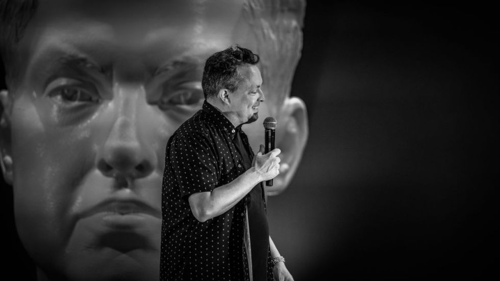 Image en noir et blanc de Mike Ward sur scène devant un buste géant de lui-même.