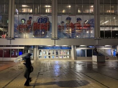 Une personne marche sous la pluie devant l'entrée principale du Centre Bell, la nuit.