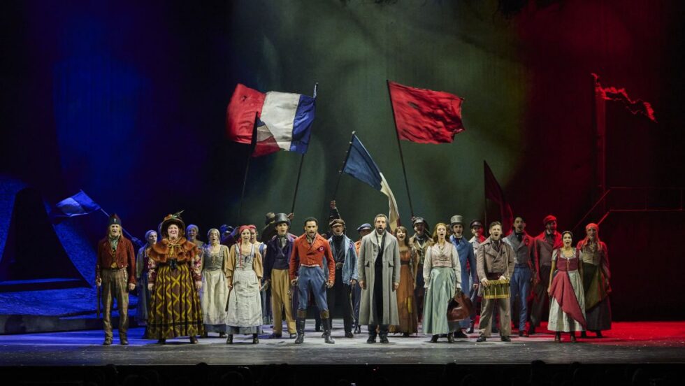 Une foule de personnages sur scène avec de grands drapeaux rouges et tricolores pendant une représentation de la comédie musicale Les Misérables.